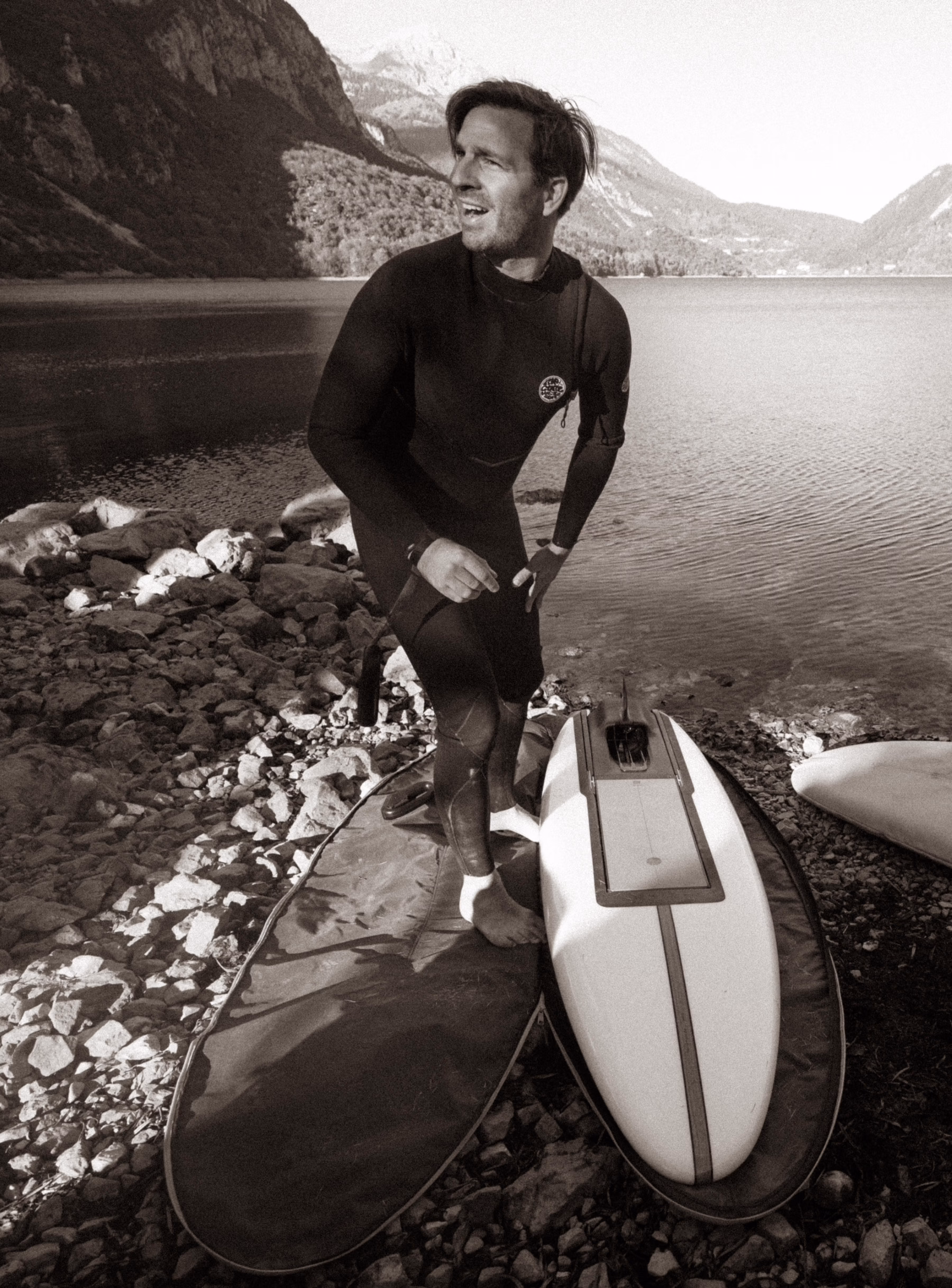 Surfer in wetsuit with two LIND electric surfboards by a mountain lake