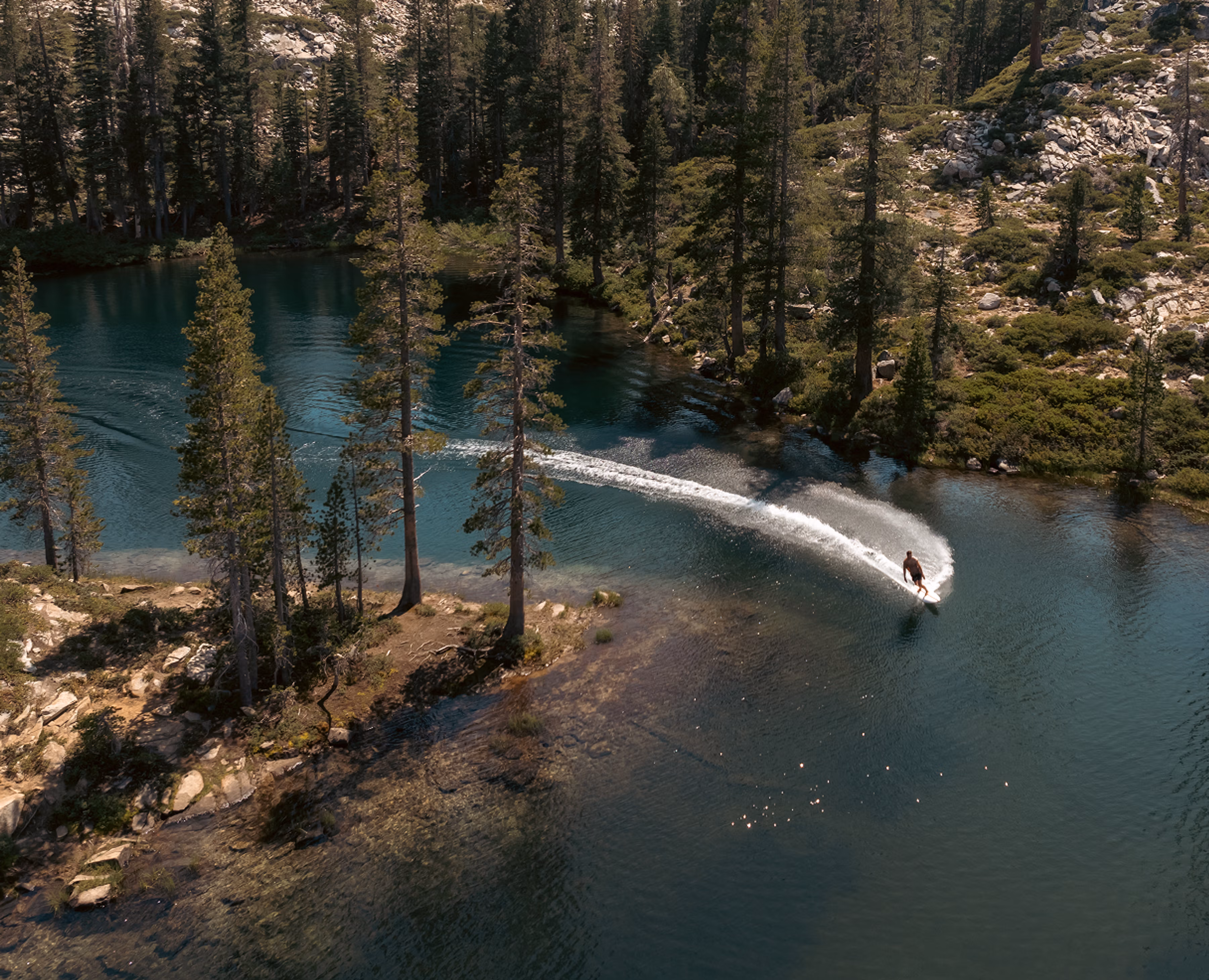 Rider on a LIND electric surfboard carving through a turquoise mountain lake, aerial view