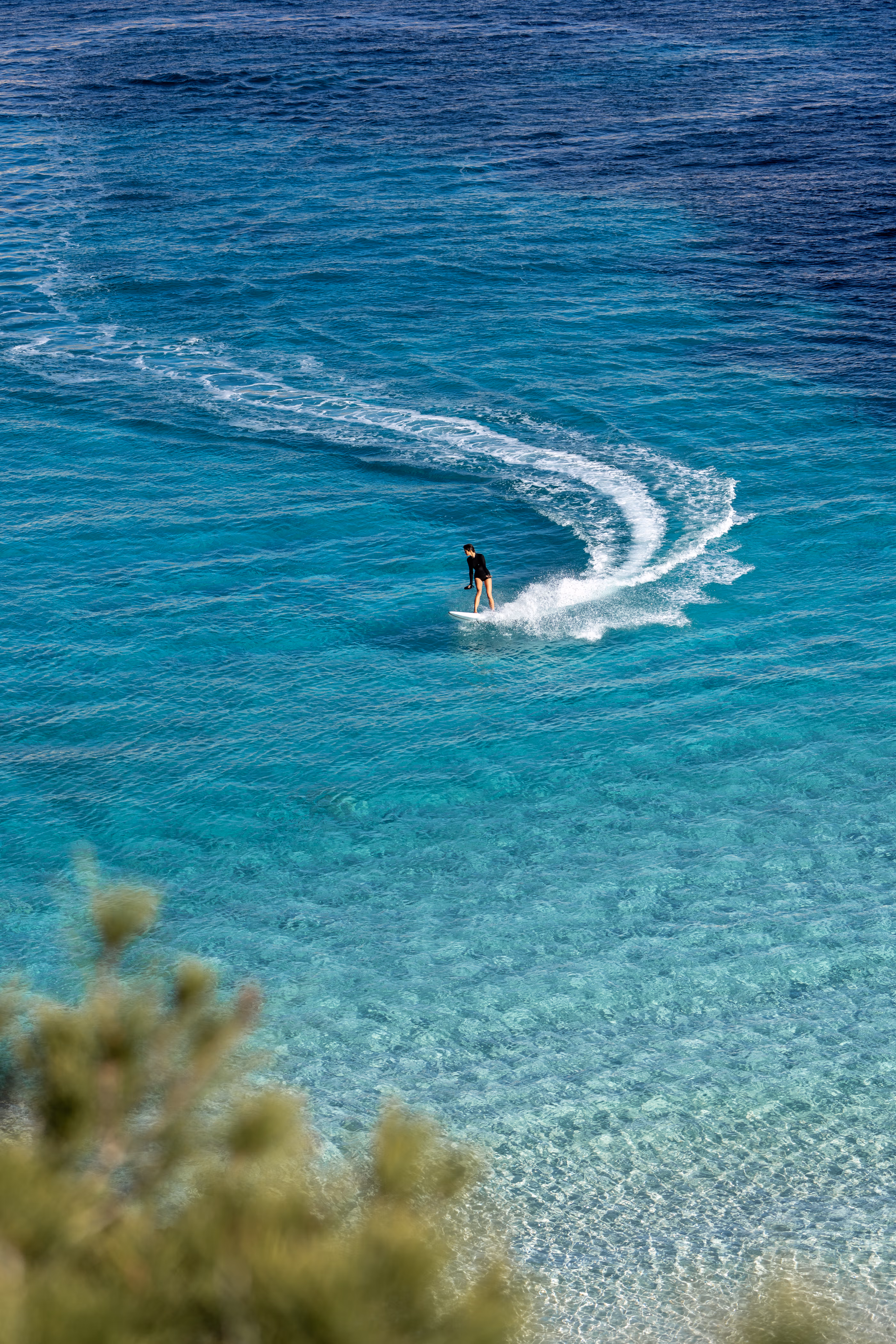 Rider on a LIND electric surfboard carving through turquoise flat water, aerial view